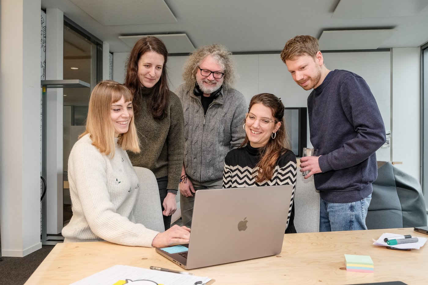 Fünf Personen aus dem Team stehen gemeinsam Um einen Laptop auf dem eine Designerin einen Entwurf zeigt. Alle wirken glücklich.