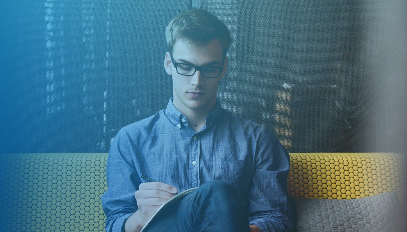 Ein Mann mit Brille sitzt auf einem Sofa und schreibt in ein Heft. Das Bild insgesamt ist mit einem blauen Filter versehen.